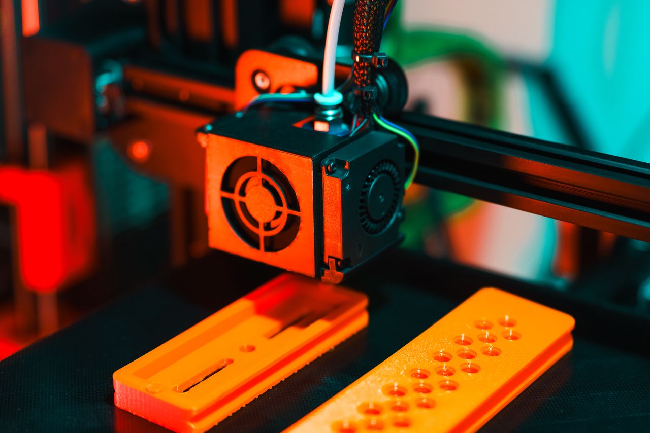 services-01 Detailed view of a 3D printer in action, extruding orange plastic components.